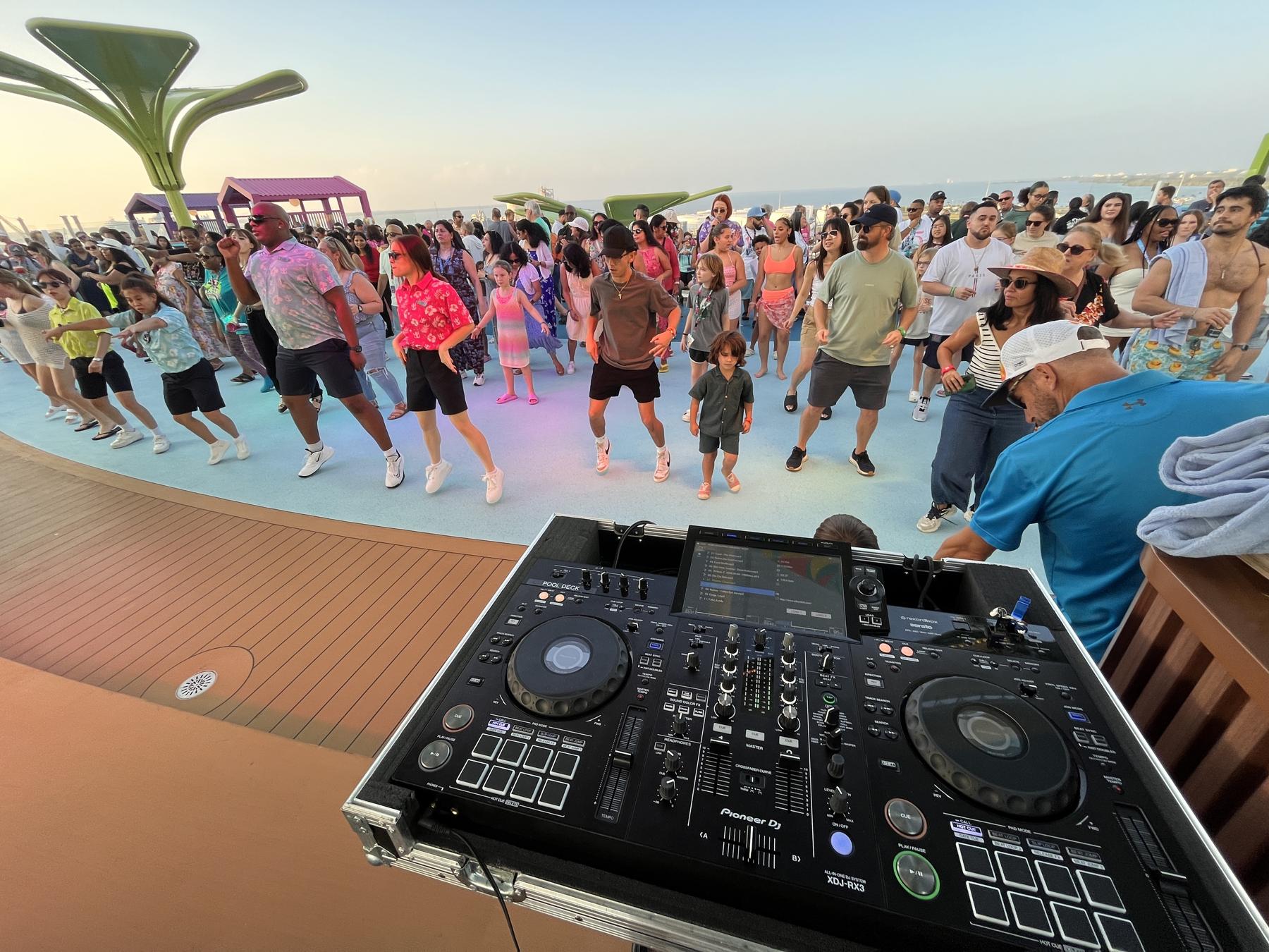 A crowd of kids and adults doing a line dance at sunset, seen from behind the DJ decks